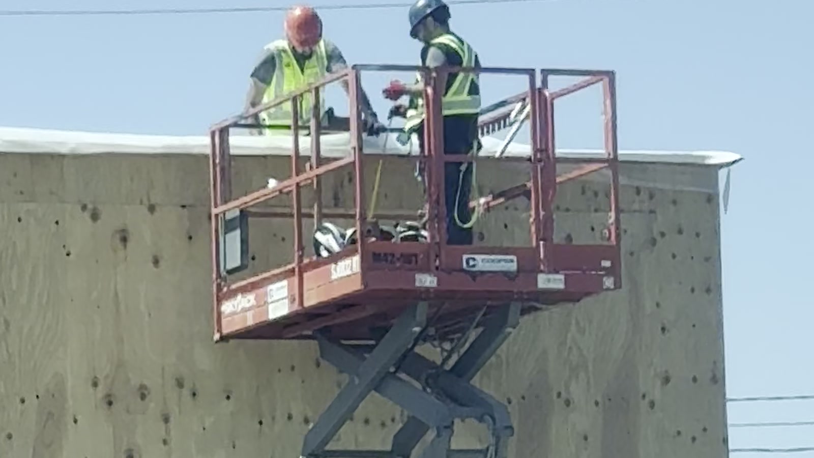 Two workers in hi-vis vests on a raised scissor lift, working at height on the edge of a building.
